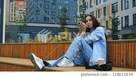 Young woman, wearing casual attire, sits on a wooden bench, savoring a takeaway coffee while engaging with her smartphone against a backdrop of modern buildings Young woman, wearing casual attire, sits on a wooden bench, savoring a takeaway coffee while engaging with her smartphone against a backdrop of modern buildings 129612674