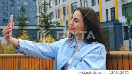 Outdoors, a young woman with long brown hair, wearing a light denim jacket, holds her smartphone and speaks into it, likely recording a voice message or using a voice assistant 129612675