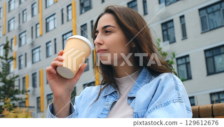 Stylish young woman sits on a bench, savoring a hot beverage from a disposable cup, taking a break amidst the backdrop of a modern building 129612676
