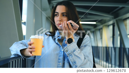 Businesswoman holding a paper cup of coffee and using her smartphone, sending voice messages and text messages and walking on a bridge in a modern city 129612678