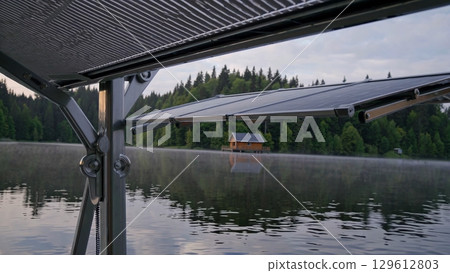 Metallic structure supporting solar panels is rotating, adjusting its angle to maximize sunlight capture, with a serene lake and a small wooden house in the background during a misty sunrise 129612803