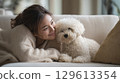 Woman cuddling with her fluffy white dog on a cozy sofa, sharing a warm smile 129613354