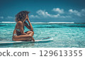 Woman sitting on surfboard in clear blue sea under sunny sky 129613355