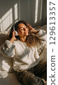 Smiling woman in cozy sweater enjoying music with headphones in warm sunlight 129613357