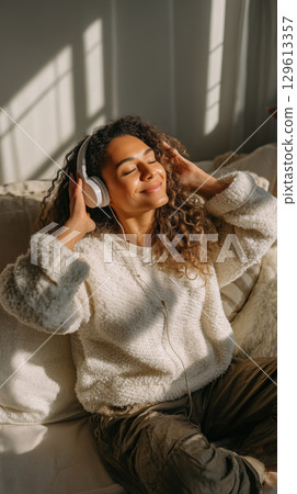 Smiling woman in cozy sweater enjoying music with headphones in warm sunlight 129613357