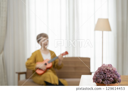 Senior woman playing the ukulele as a hobby on the sofa in the living room Senior woman playing the ukulele as a hobby on the sofa in the living room 129614453