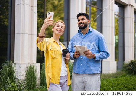 A diverse couple taking a selfie outdoors with a smartphone and tablet in front of a modern building. 129614553
