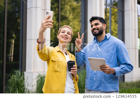 A woman takes a selfie with a phone, while a man makes a peace sign and holds a tablet outdoors. They are smiling. 129614555