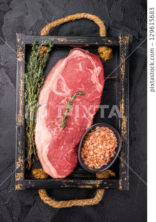 Raw New York strip beef steak in a wooden tray with herbs. black background. top view 129615683