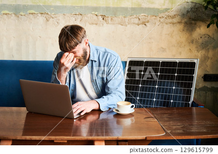Bearded man works on laptop at wooden table, with cup of coffee. Solar panel nearby, emphasizing sustainable eco-friendly workspace that combines technology and sustainability in cozy environment. Bearded man works on laptop at wooden table, with cup of coffee. Solar panel nearby, emphasizing sustainable eco-friendly workspace that combines technology and sustainability in cozy environment. 129615799