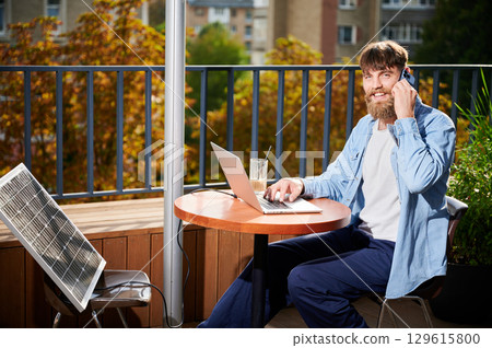 Man works on laptop and smartphone at outdoor table, with glass of iced coffee. Solar panel nearby, emphasizing sustainable, modern eco-friendly workspace that combines technology and sustainability. 129615800