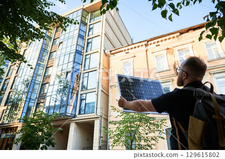 Man holding photovoltaic solar panel in front of new building. Concept of integration of sustainable renewable energy sources into modern architecture. 129615802