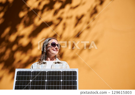 Woman holding solar panel for charging, stands against vibrant orange wall. Smiling girl wears sunglasses, white top. Artistic shadows cast by nearby trees branches on wall. 129615803