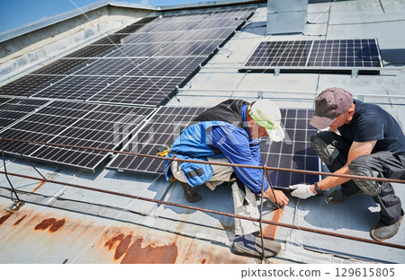 Workers building solar panel system on metal rooftop of house. Two men installers installing photovoltaic solar module outdoors. Alternative, green and renewable energy generation concept. Workers building solar panel system on metal rooftop of house. Two men installers installing photovoltaic solar module outdoors. Alternative, green and renewable energy generation concept. 129615805