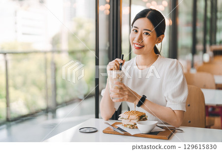 Happy asian woman healthy eating breakfast sandwich at cafe sitting near window indoors on day Happy asian woman healthy eating breakfast sandwich at cafe sitting near window indoors on day 129615830