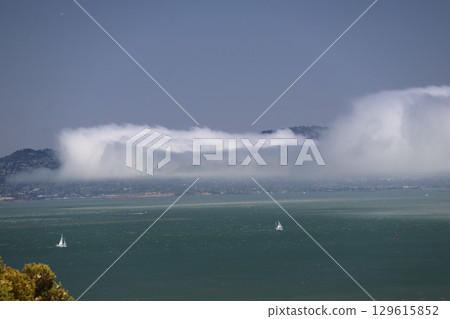 A view of the blue sky, the sea with white sail yachts, and the San Francisco cityscape with misty clouds. 129615852