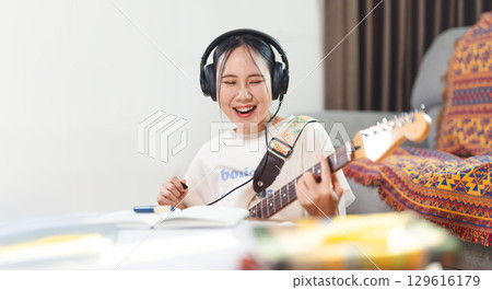 Young adult asian woman playing electric guitar music wearing headphones enjoy hobby at home 129616179