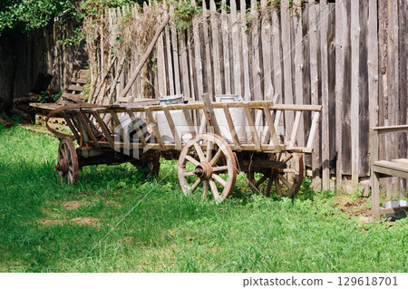 Old wooden cart sits on green grass in a rural setting with a fence backdrop during the daytime Old wooden cart sits on green grass in a rural setting with a fence backdrop during the daytime 129618701