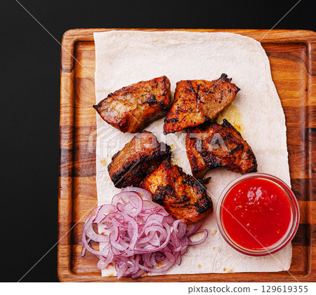 Juicy grilled meat served with onions and spicy sauce on a wooden tray 129619355