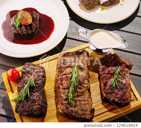 Grilled steaks arranged beautifully on a wooden platter with sauce and garnishes 129619924