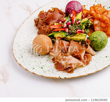 A gourmet platter with cured meat slices, colorful cheese balls, crispy chips, avocado topped with pesto, chili, and pomegranate seeds A gourmet platter with cured meat slices, colorful cheese balls, crispy chips, avocado topped with pesto, chili, and pomegranate seeds 129620090