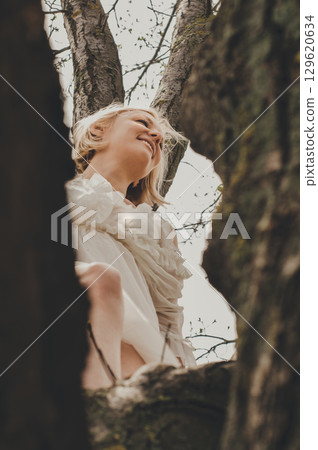 a beautiful young woman in a white chiffon dress with a short blonde haircut is sitting on a tree, bottom view 129620634