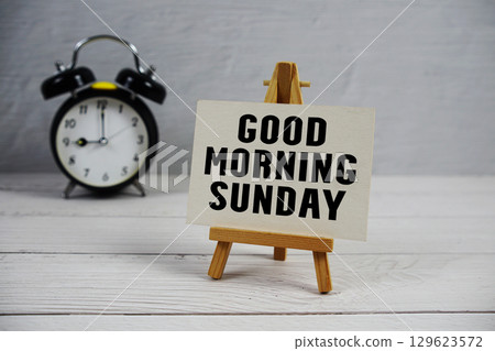 Good Morning Sunday text with easel and alarm clock on wooden background Good Morning Sunday text with easel and alarm clock on wooden background 129623572