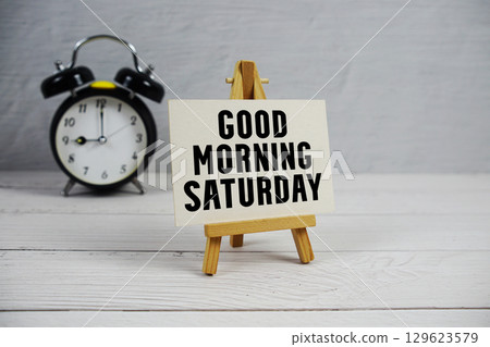 Good Morning Saturday text with easel and alarm clock on wooden background 129623579