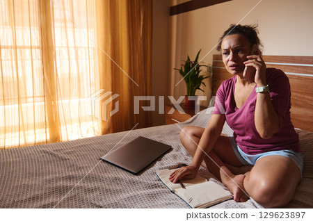 Concerned Woman Talking on Phone While Sitting on Bed at Home 129623897