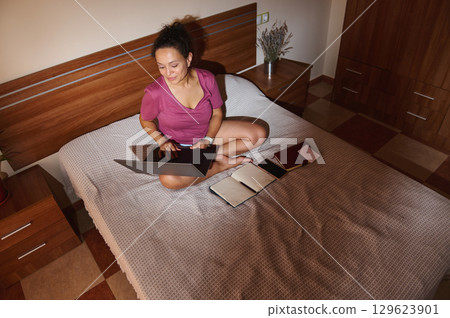 Woman Using Laptop on Bed with Notes and Journals Around Her 129623901
