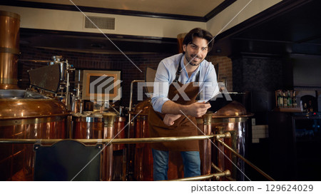 A man wearing an apron is studying a clipboard in a brewery setting 129624029