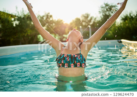 Teen girl enjoying a summer day in a pool, celebrating with arms raised under the warm sun 129624470