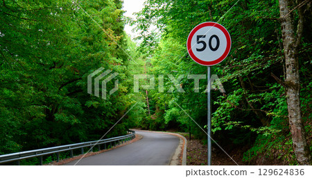 speed limit sign stands near a forested road, surrounded by lush greenery speed limit sign stands near a forested road, surrounded by lush greenery 129624836