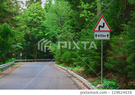 A winding road sign stands beside a winding path in a lush green forest A winding road sign stands beside a winding path in a lush green forest 129624838