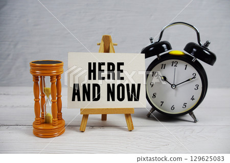 Here And Now text with hourglass and alarm clock on wooden background 129625083