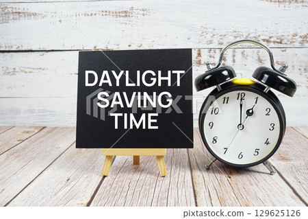 Daylight Saving Time text with easel and alarm clock on wooden background 129625126