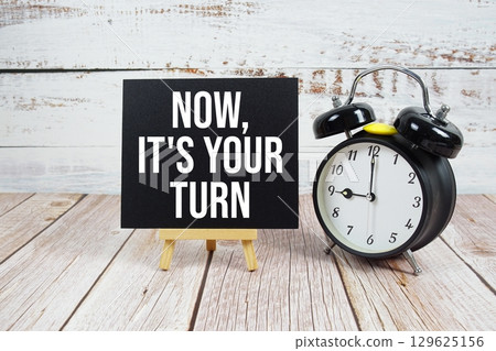 Now, it's Your Turn text with easel and alarm clock on wooden background 129625156
