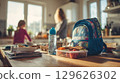 Mother preparing school backpack with lunchbox and books in kitchen 129626302