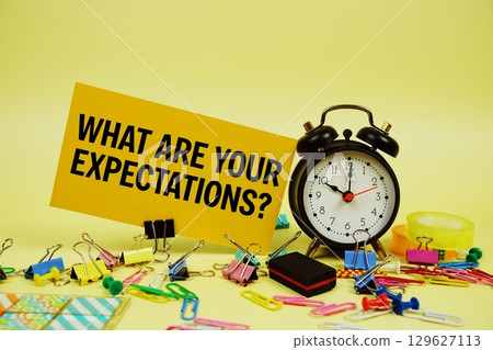 What Are Your Expectations? text on paper card with alarm clock and stationery on yellow background What Are Your Expectations? text on paper card with alarm clock and stationery on yellow background 129627113