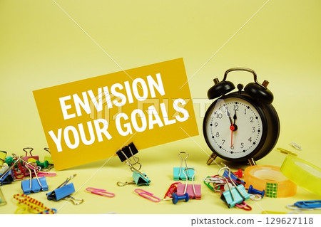 Envision Your Goals text on paper card with stationery and alarm clock on yellow background 129627118