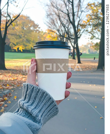 Woman Holding White Coffee Cup on a Sidewalk with Colorful Autumn Trees Background 129627988
