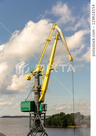 A tall yellow port crane against a blue sky with white clouds, on the shore of a bay 129628554