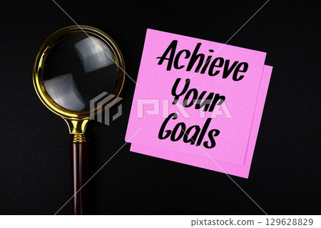 Achieve Your Goals text on sticky note with magnifying glass on black background 129628829