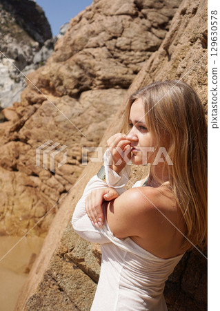 A Serene Beach Model Posed Gracefully by the Rocks in a Beautiful Sunlit Landscape 129630578