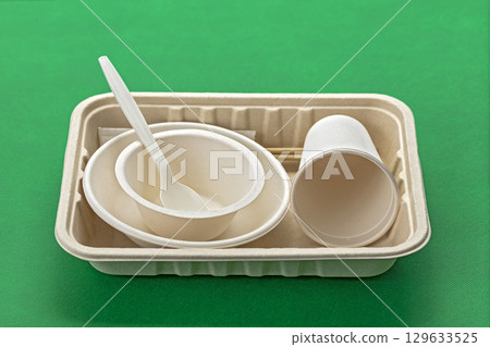 Biodegradable disposable dishes including bowls, cup, and cutlery on green background, showcasing eco friendly tableware options 129633525