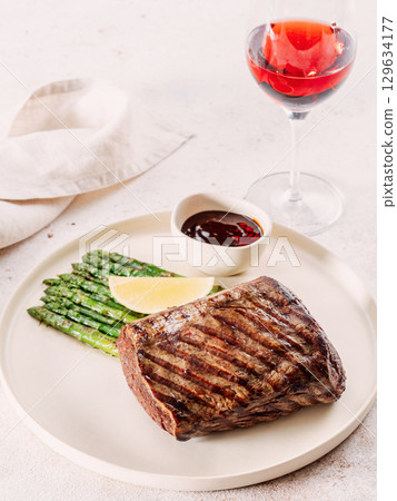 Grilled meat steak on plate with lemon and asparagus on white table background. Food and cuisine concept. Restaurant-style plating, with red wine glass on table 129634177