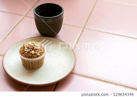 cup of coffee and muffin sit on a white plate on pink tiled table. muffin is sprinkled with nuts. cup of coffee and muffin sit on a white plate on pink tiled table. muffin is sprinkled with nuts. 129634346