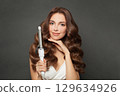 Lovely young model woman with long curly wavy hairstyle showing hot curling tongs iron 129634926