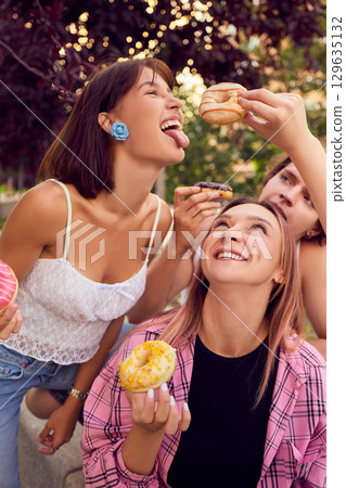 Close up of friends smiling and offering colorful donuts outdoors 129635132