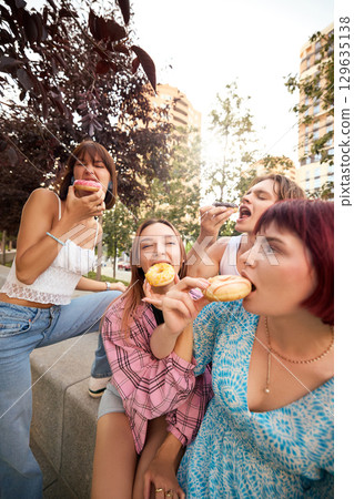Playful young friends eating donuts and laughing together in urban outdoor setting 129635138
